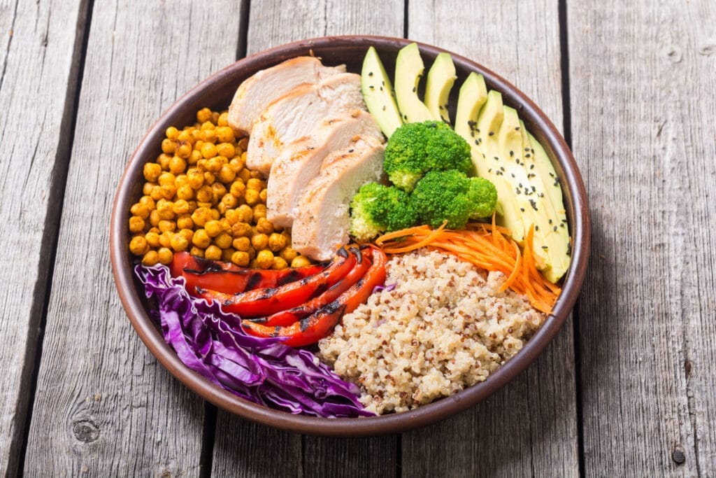 Buddha bowl with avocado, chicken, peppers, quinoa, chickpeas, and other vegetables on wooden background.