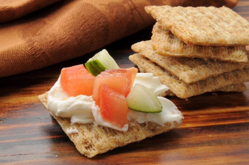 A whole wheat cracker topped with cream cheese, tomato, cucumber, and chives, with other whole wheat crackers in the background on a dark brown surface.